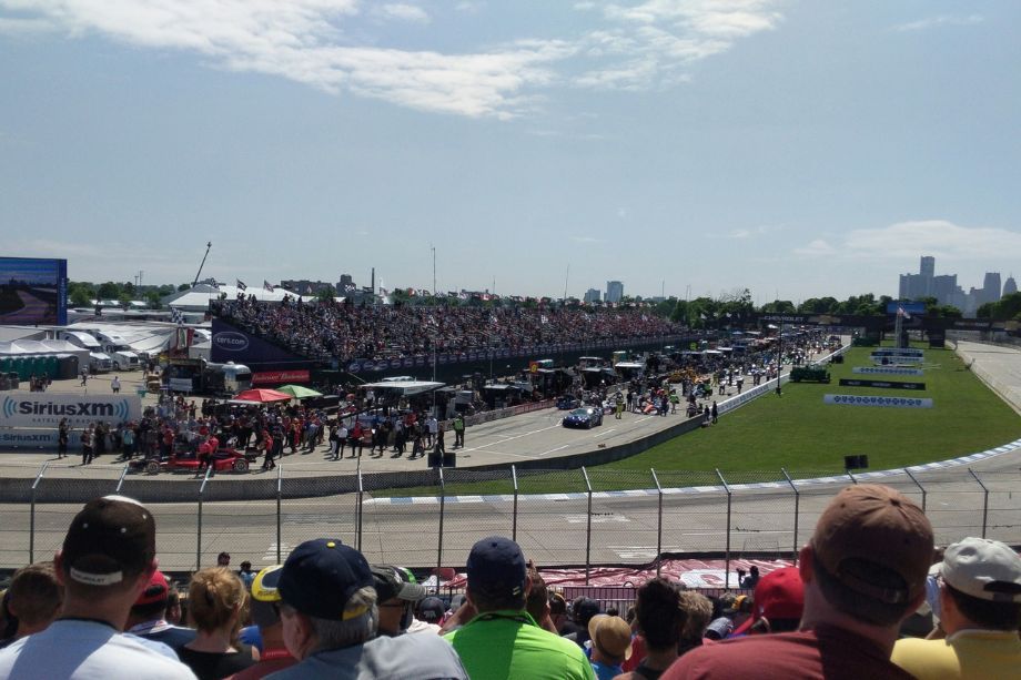 Crowd watches a car race from bleachers and pit area along a sunny oval track with a distant city skyline.